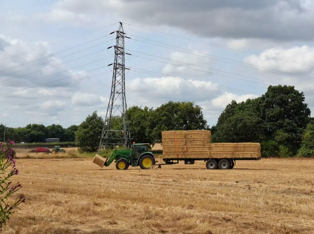 A green tractor with a straw bale attached to prongs at the front pulls a trailer loaded with straw bales