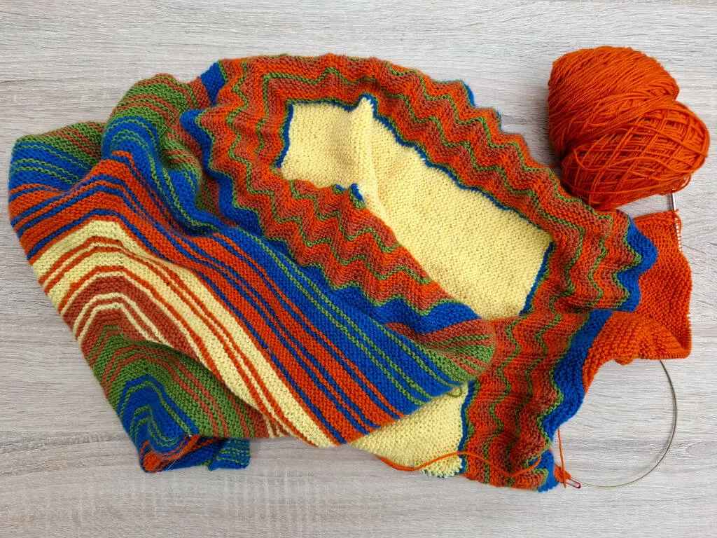 A partly-knitted shawl in yellow, green, pumpkin, orange and blue yarn lies on a wooden table 