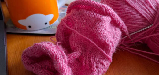 A tube of knitting in pink yarn lying on a wooden table next to an orange mug and an open book