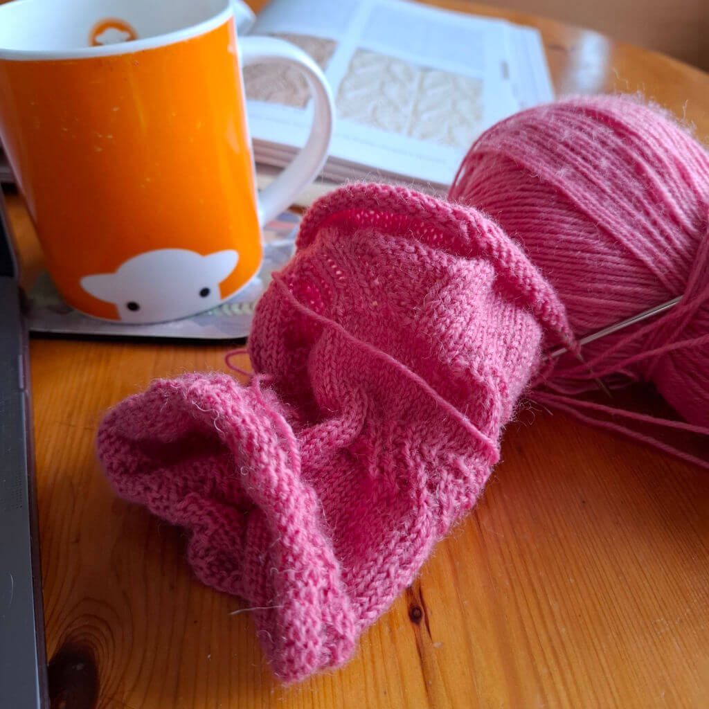 A tube of knitting in pink yarn lying on a wooden table next to an orange mug and an open book