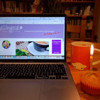 An open laptop with the latest Winwick Mum blog post on the screen. Next to the laptop on the wooden table is an orange mug of tea and a cupcake with a candle in it. In the background are bookcases filled with books