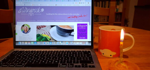 An open laptop with the latest Winwick Mum blog post on the screen. Next to the laptop on the wooden table is an orange mug of tea and a cupcake with a candle in it. In the background are bookcases filled with books