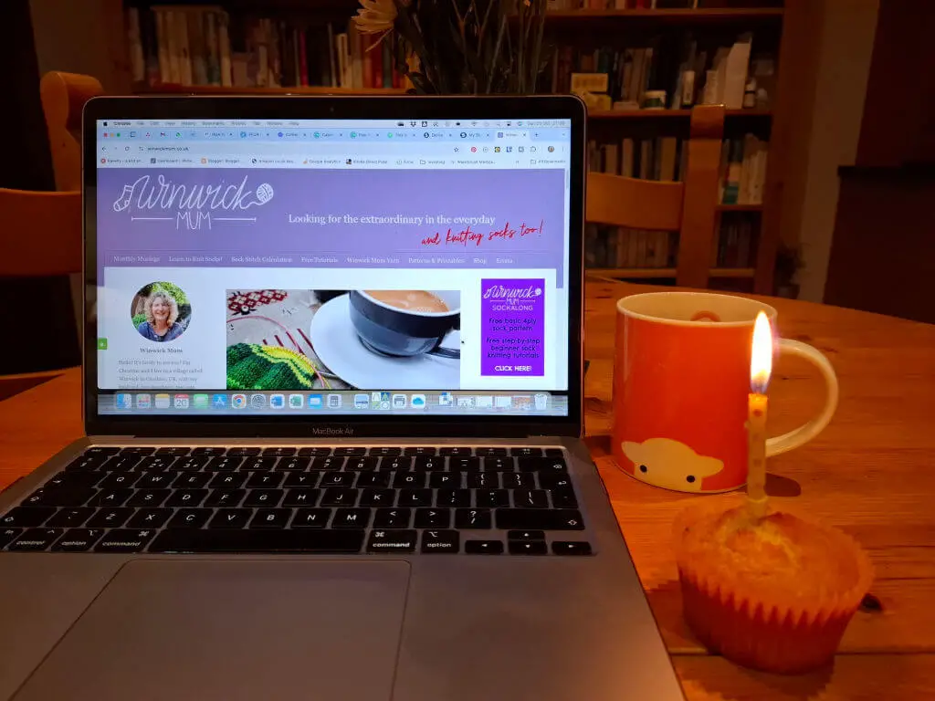 An open laptop with the latest Winwick Mum blog post on the screen. Next to the laptop on the wooden table is an orange mug of tea and a cupcake with a candle in it. In the background are bookcases filled with books