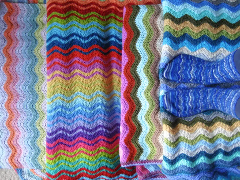 Four Attic24 Neat Ripple blankets arranged to show off the stripes, with Lucy standing on one of the blankets wearing a pair of Winwick Mum Neat Ripple socks in blue striped yarn