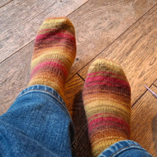A pair of socks in stripes of brown, red, green and gold on a pair of feet. The person wearing them is wearing jeans and the feet are resting on a wooden floor