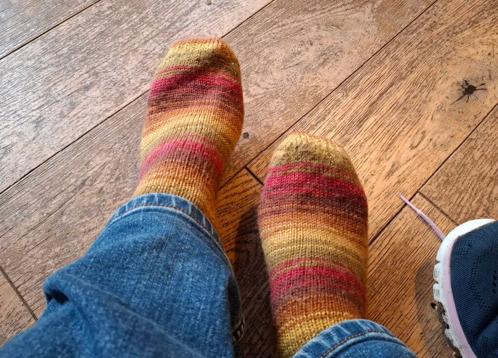 A pair of socks in stripes of brown, red, green and gold on a pair of feet.  The person wearing them is wearing jeans and the feet are resting on a wooden floor
