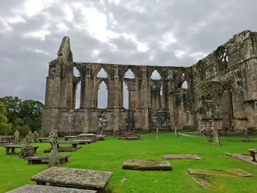 Stone ruins of a monastery against a grey cloudy sky.  There is a grassy area with old gravestones in the foreground
