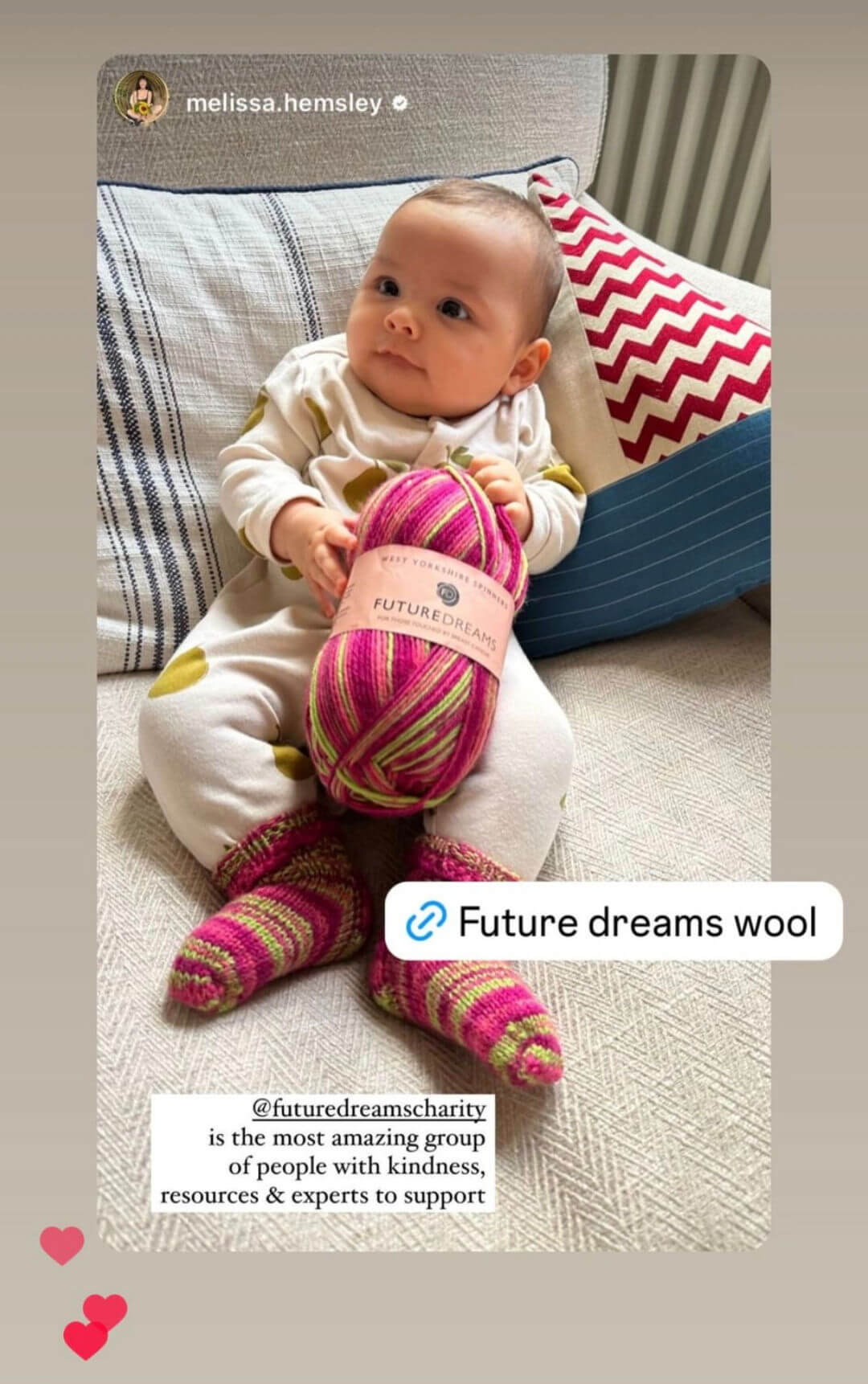 A baby in a white babygro leaning against a striped cushion wears a tiny pair of hand knitted socks and holds a ball of pink striped yarn