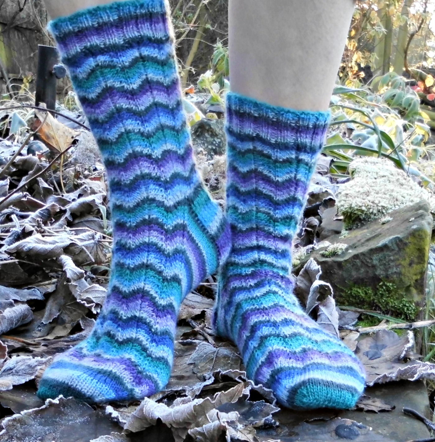 A pair of striped socks in shades of blue which ripple around the sock. The socks are modelled on feet and the model is standing on large stones