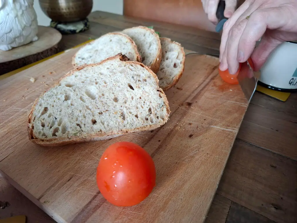 Slices of home made bread on a wooden chopping board.  There is a tomato on the board, and a pair of man's hands cutting another tomato with a knife