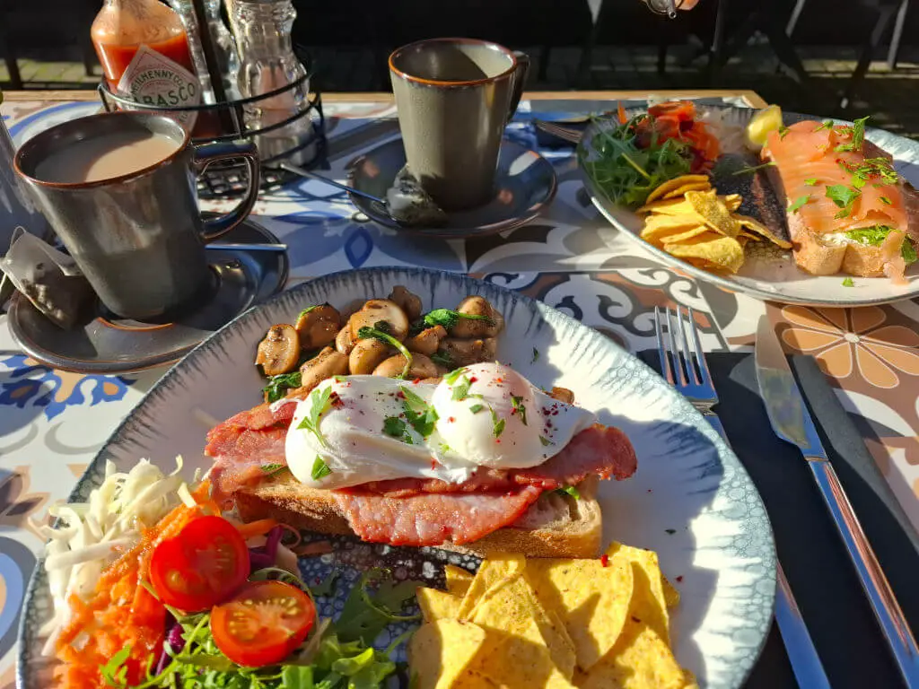 A plate of poached eggs on toast with bacon and mushrooms with salad and tortilla chips on an outdoor restaurant table. There is a second plate of food to the right, and two mugs for hot drinks on the table