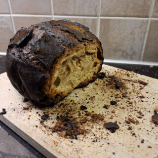 A burnt loaf with the end cut off sits on a chopping board surrounded by crumbs