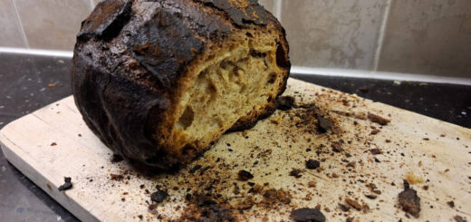 A burnt loaf with the end cut off sits on a chopping board surrounded by crumbs