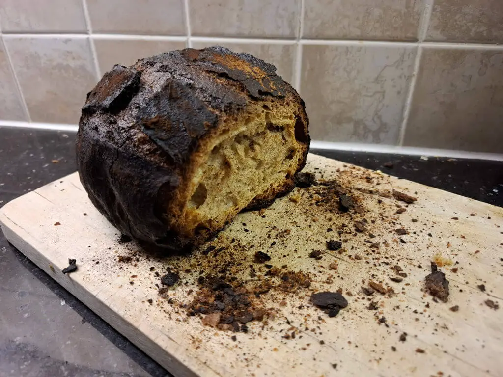 A burnt loaf with the end cut off sits on a chopping board surrounded by crumbs