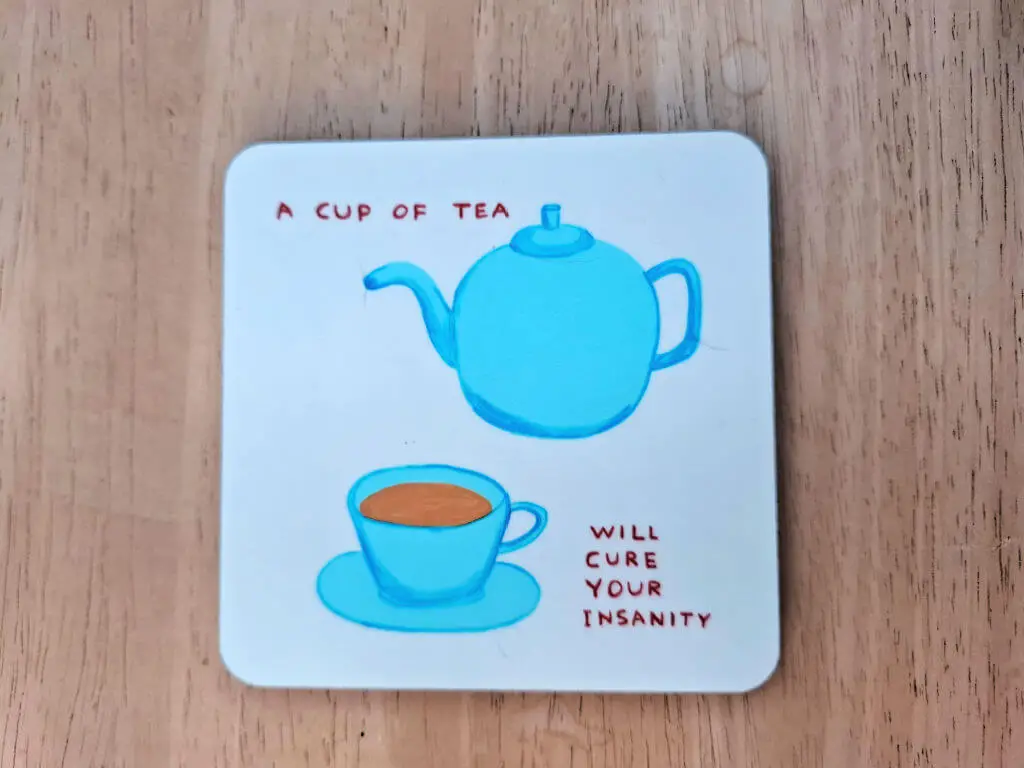 A square white coaster with rounded edges, with a picture of a blue teapot and cup and saucer on it. The words on the coaster are "A cup of tea will cure your insanity"