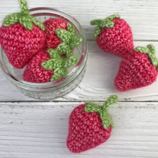 A glass jar on a white wooden painted board containing crochet strawberries