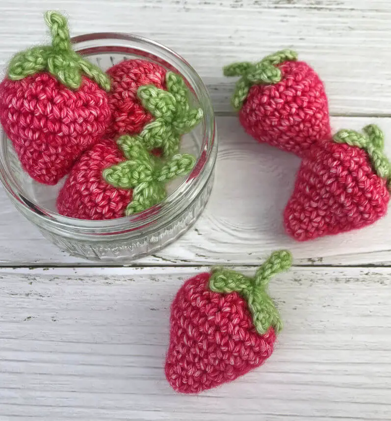 A glass jar on a white wooden painted board containing crochet strawberries
