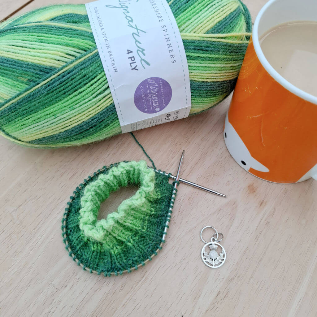 A knitted sock cuff in green striped yarn on a circular needle. On the table next to the knitting is a stitch marker in the shape of a thistle, an orange mug of tea and a ball of green striped yarn