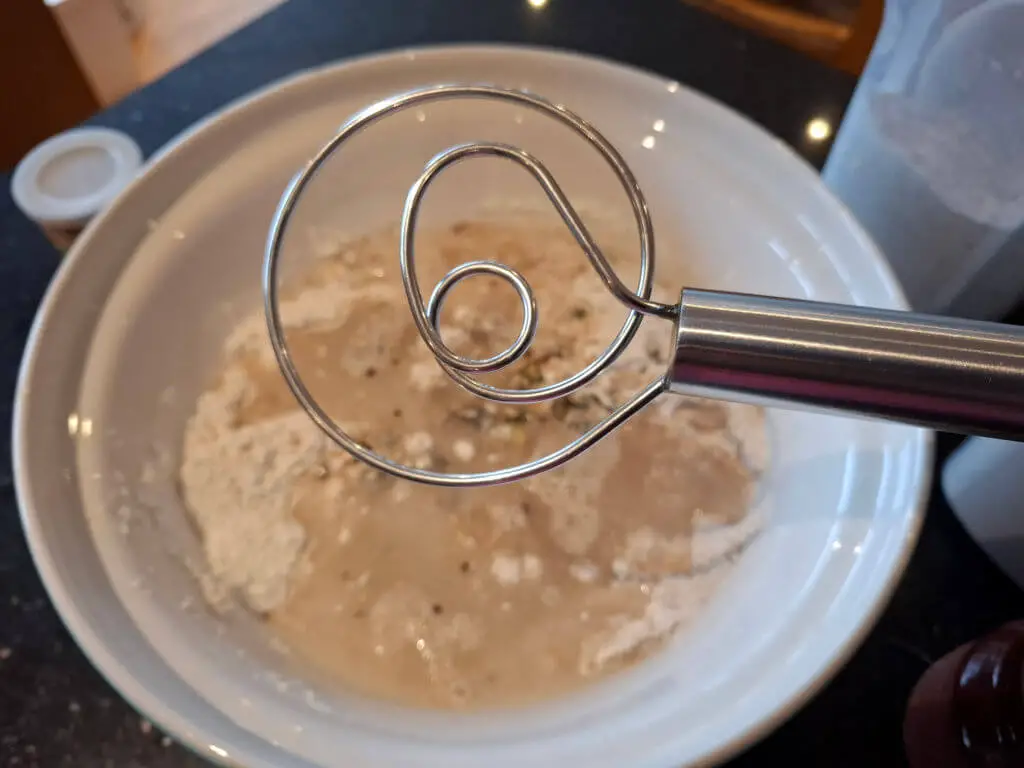 A metal whisk with a particular curled shape held over a mixing bowl of flour