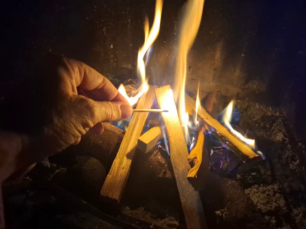 A hand holding a match to start a fire. There are already flames around the kindling