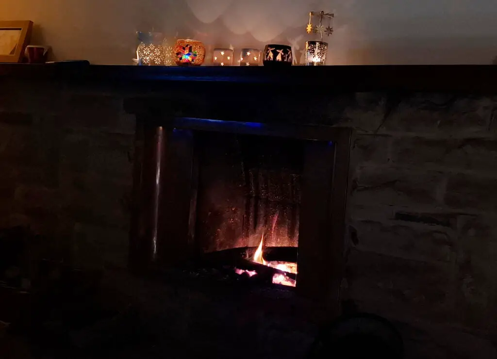A fire burns in an open fireplace. On the mantelpiece above are glowing tealights in various holders