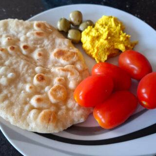 A flatbread on a black and white plate with red plum tomatoes, green olives and golden hummus