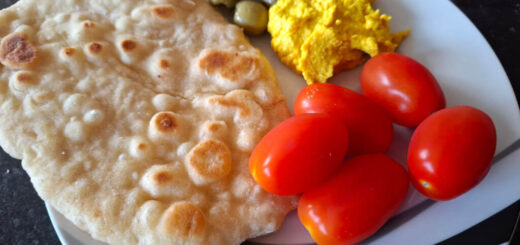 A flatbread on a black and white plate with red plum tomatoes, green olives and golden hummus