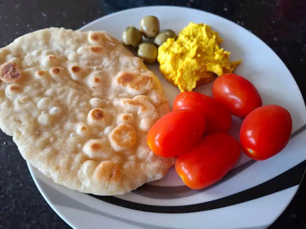 A flatbread on a black and white plate with red plum tomatoes, green olives and golden hummus