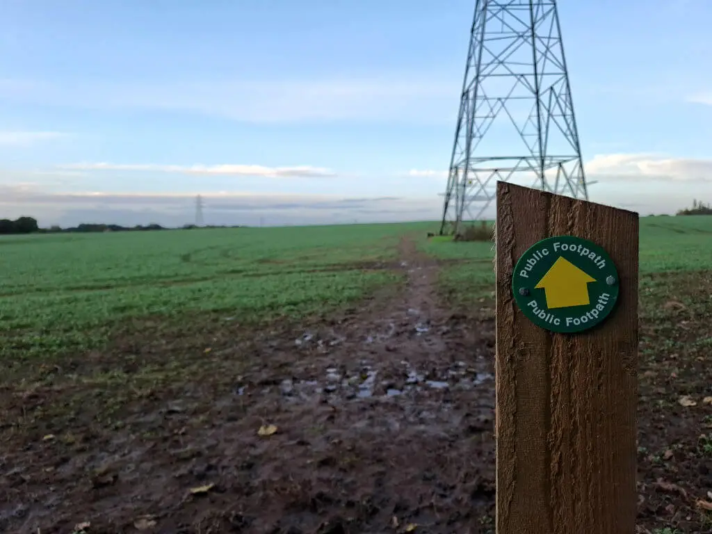 A circular footpath direction sign on a wooden post with a muddy footpath across a green field in the background