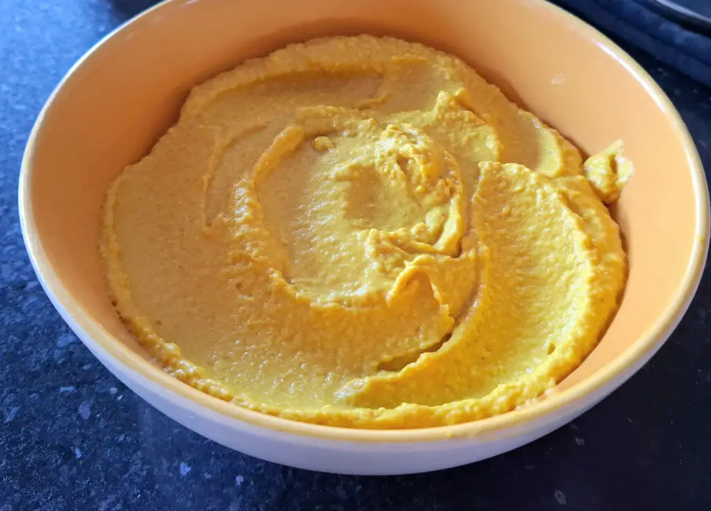 A yellow bowl on a black granite worktop, filled with golden-coloured hummus