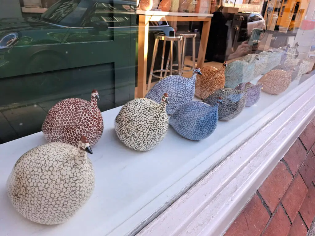 A line of pottery guinea hens in a shop window. They are shades of blue, stone, grey and terracotta