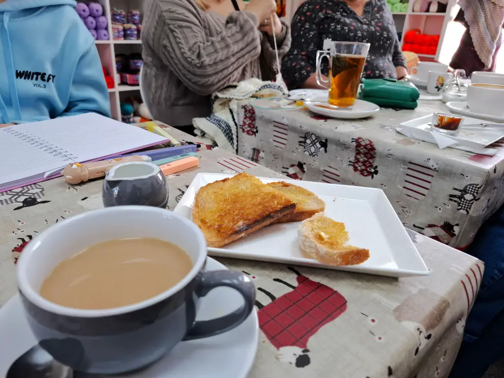 Two tables in a cafe with cups of tea and coffee on them and  a plate of toast.  There are people sitting around the tables, one of them is working on a knitted jumper