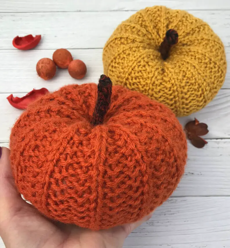 Two knitted pumpkins against a white painted board background. One pumpkin is orange and is held in someone's hand, the other is golden and is sitting on the board