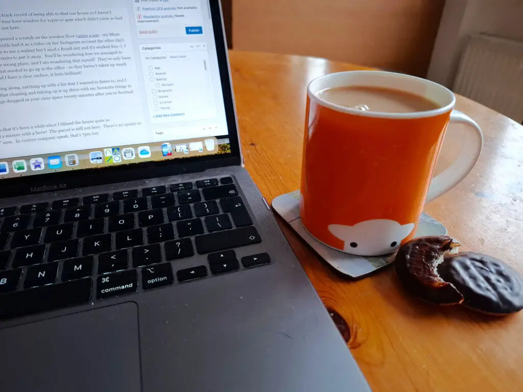An orange mug of tea on a wooden table next to a laptop and two chocolate biscuits