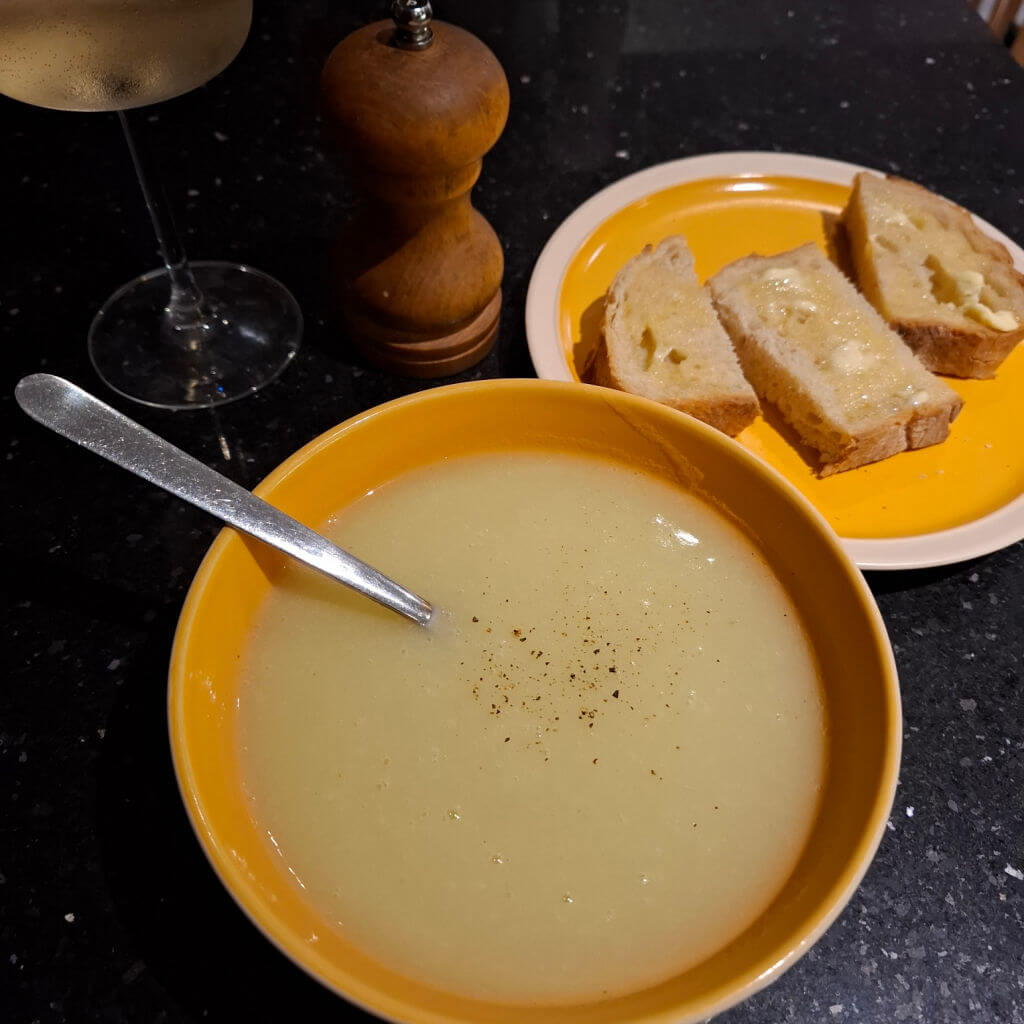 A bowl of pale green soup next to a yellow plate with sliced bread on it.  There is also a pepper grinder and a wine glass next to the bowl