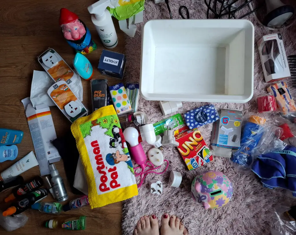 A big pile of travel plugs, sun cream bottles, face flannels and other toiletries on a pink furry rug next to an empty white storage box