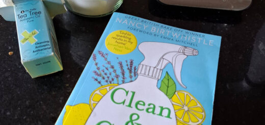 A copy of a book with a blue, green and yellow cover entitled "Clean & Green" lies on a granite worktop next to a bottle of tea tree oil, a jar of citric acid and a measuring jug on kitchen scales.
