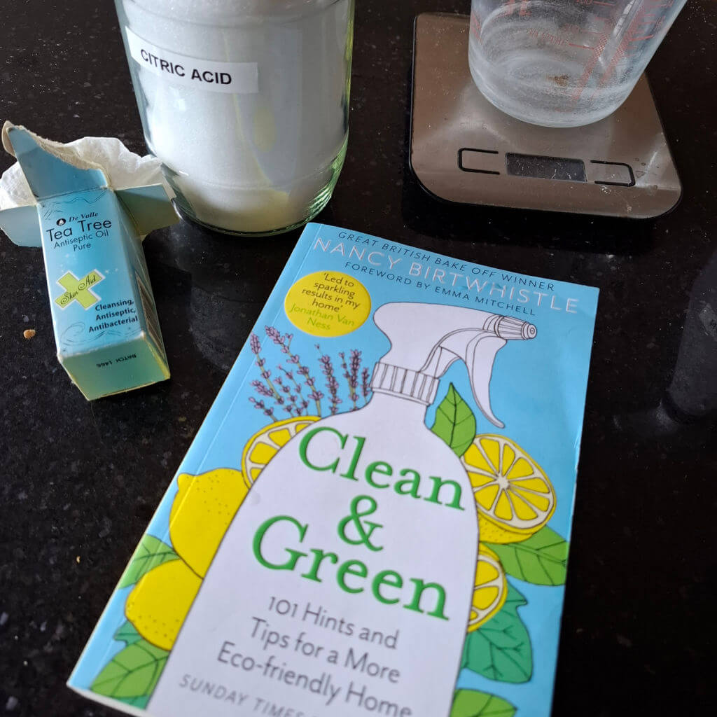 A copy of a book with a blue, green and yellow cover entitled "Clean & Green" lies on a granite worktop next to a bottle of tea tree oil, a jar of citric acid and a measuring jug on kitchen scales.