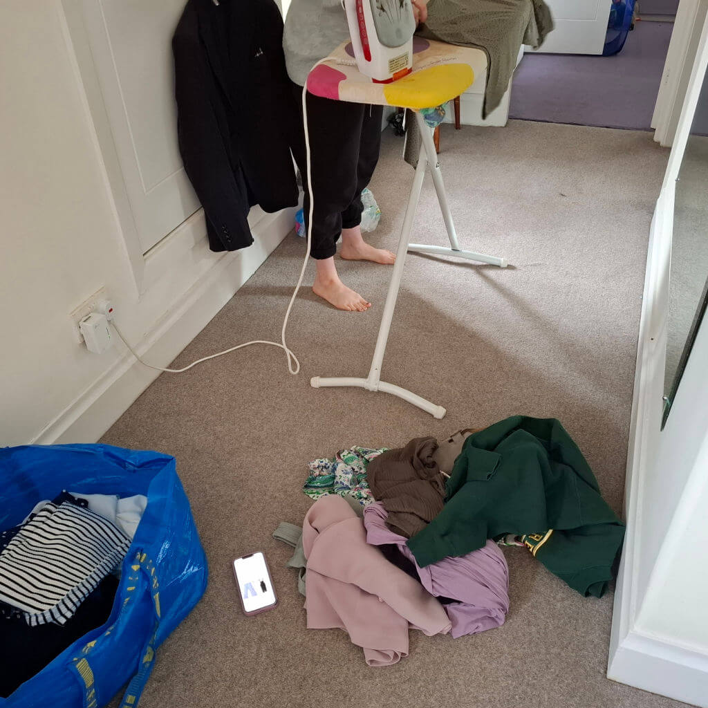 A girl in black joggers stands at an ironing board in a hallway. There are bags of clothes on the floor