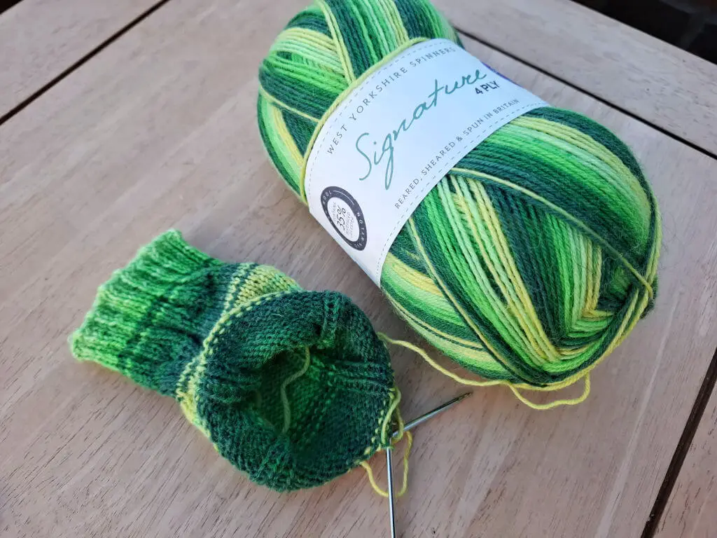 A partly-knitted green striped sock on a wooden table next to the ball of yarn