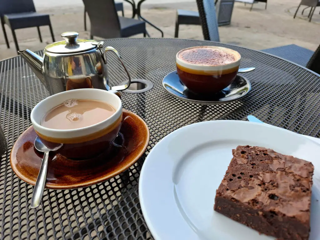 A cup of tea in a saucer on a metal outdoor table, next to a cup of frothy cappucino and a slice of chocolate brownie on a white plate