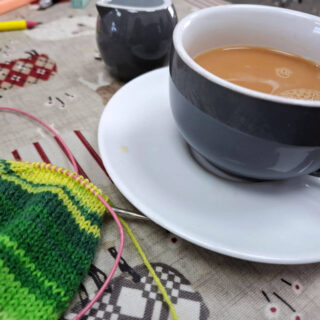 A blue teacup with a white inside on a white saucer sitting on a table next to a partly-knitted green striped sock