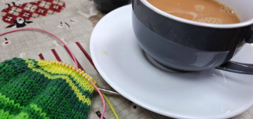 A blue teacup with a white inside on a white saucer sitting on a table next to a partly-knitted green striped sock