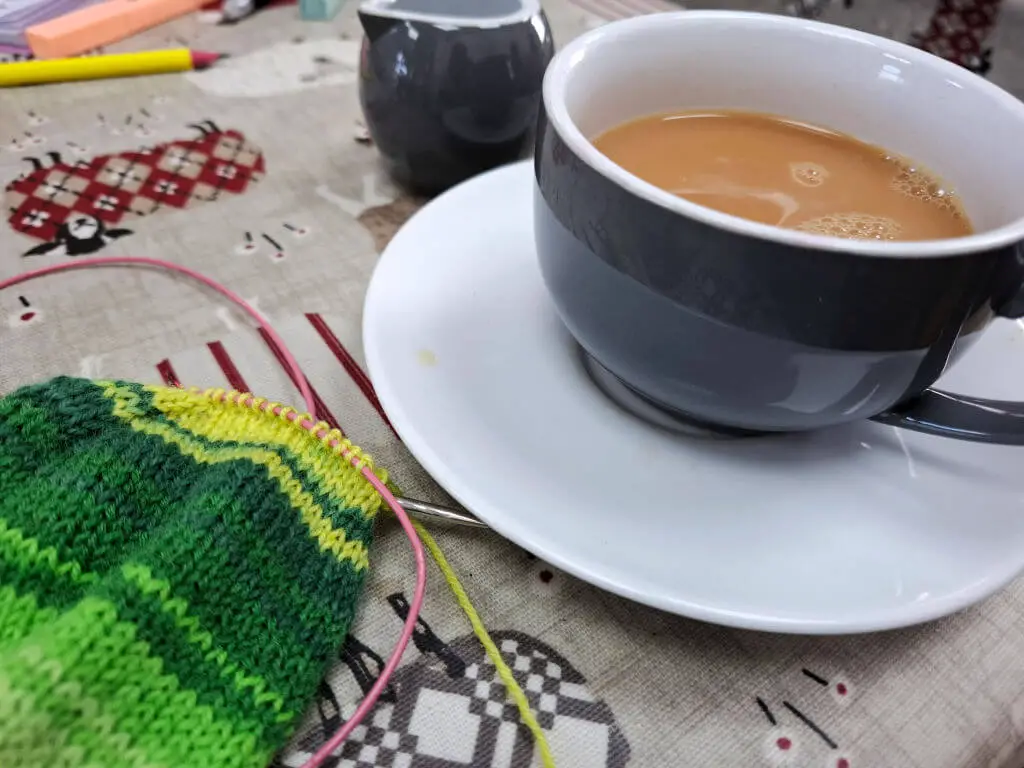 A blue teacup with a white inside on a white saucer sitting on a table next to a partly-knitted green striped sock