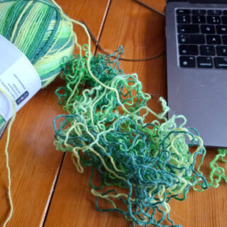 Unravelled green yarn on a wooden table next to an open laptop and the rest of the ball of yarn