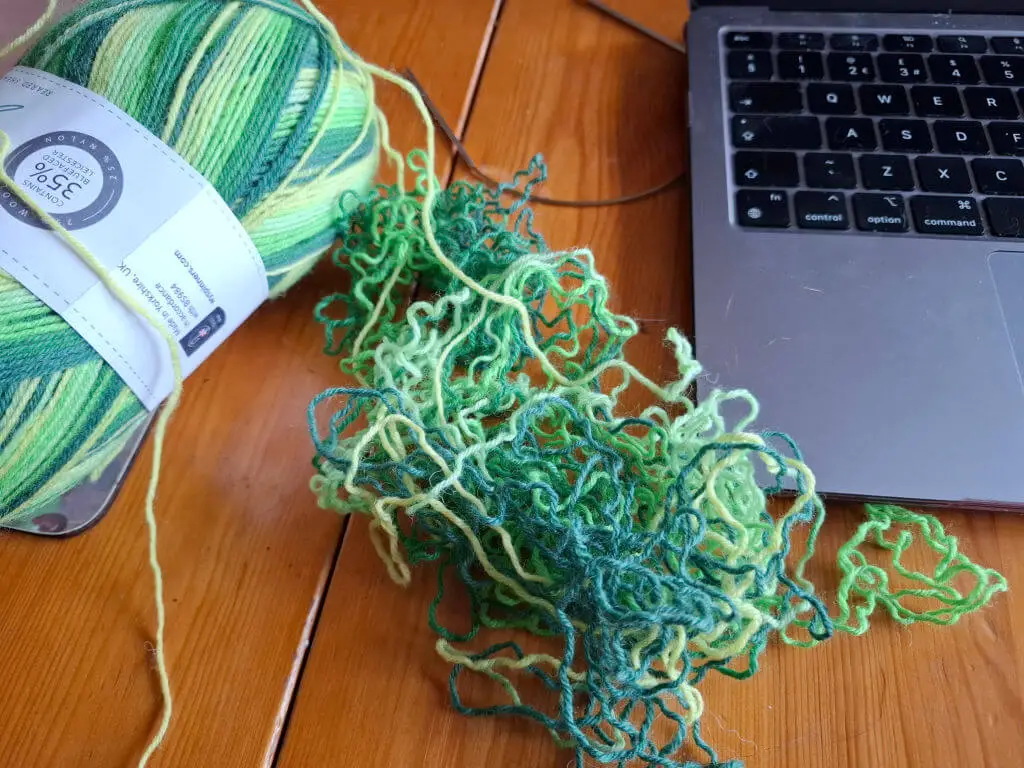 Unravelled green yarn on a wooden table next to an open laptop and the rest of the ball of yarn
