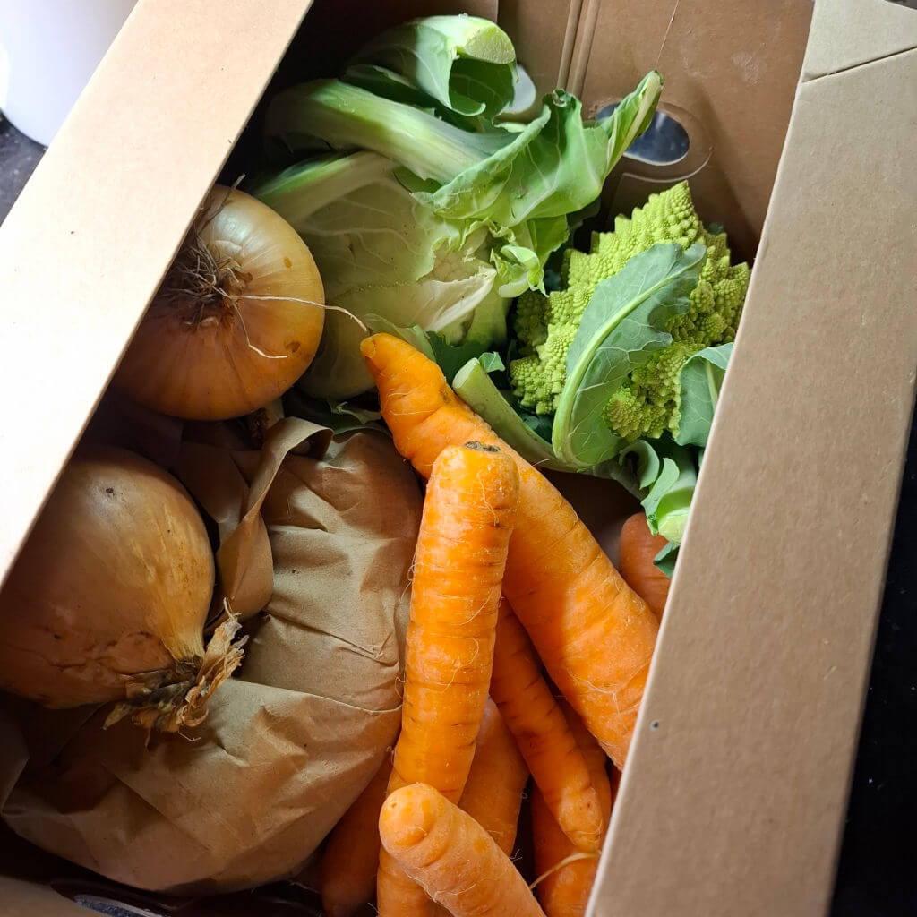 A brown cardboard box containing a paper bag of potatoes, carrots, onions, cauliflower and Romanesco 