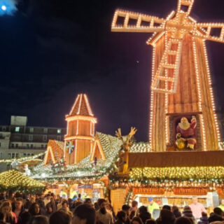 A view across a busy square to Christmas market stalls light up with lights. A huge windmill is on the roof of the stalls.