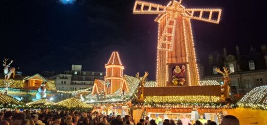 A view across a busy square to Christmas market stalls light up with lights. A huge windmill is on the roof of the stalls.