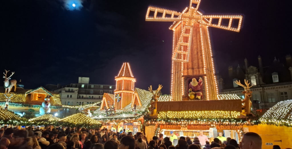 A view across a busy square to Christmas market stalls light up with lights.  A huge windmill is on the roof of the stalls.